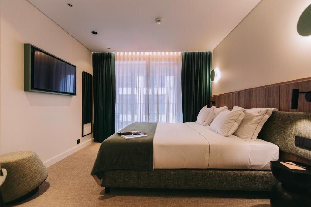 A modern hotel room with a large bed, a flat-screen TV mounted on the wall, and a window with sheer curtains.