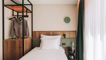 Une chambre d’hôtel avec un lit, une tête de lit en bois, un porte-manteau mural, un élément de décoration circulaire fixé au mur, et une table de chevet avec une lampe et une bouteille.