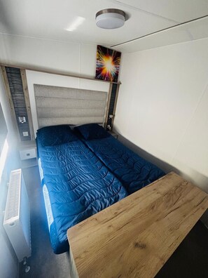 A compact bedroom with a bed, a headboard, a wooden bench, a wall-mounted shelf, and a ceiling light fixture.