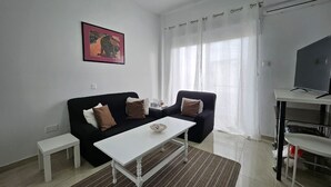 A modern living room with a black sofa, a white coffee table, a small white side table, a TV on a stand, and a window with sheer curtains. - Larnaca Sunset One-bedroom Suite Close to the Beach (Larnaca)