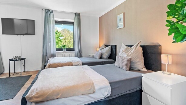 A hotel room with two beds, a flat-screen TV, a small table with a 'LOVE' sign, a window with curtains, and a potted plant. - Oasis by Loftigo - Cozy Apartment with Rain Shower (Selfkant)