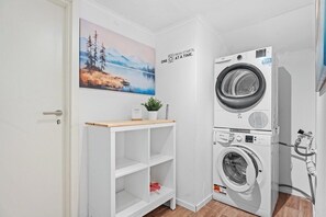 A laundry room with a stacked washer and dryer, a white shelving unit, a painting on the wall, and a door.