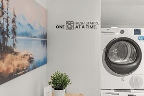 A laundry area with a washing machine, a wall-mounted sign that reads 'FRESH STARTS, ONE AT A TIME,' and a framed landscape painting. - Forest by Loftigo - Patio & Sunroom (Selfkant)