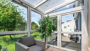 Quarto iluminado pelo sol, com um sofá de vime, uma planta em vaso e vista para uma casa branca através de grandes portas de vidro.