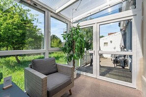 Een zonnige kamer met een rieten bank, een potplant en uitzicht op een wit huis door grote glazen deuren.