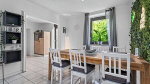 Cozinha moderna com mesa de madeira e cadeiras brancas, prateleira embutida com plantas e vista para o verde lá fora.