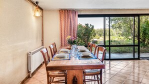 Un coin repas avec une longue table en bois, dressée pour quatre personnes, entourée de chaises, et offrant une vue sur la verdure à travers une porte vitrée.