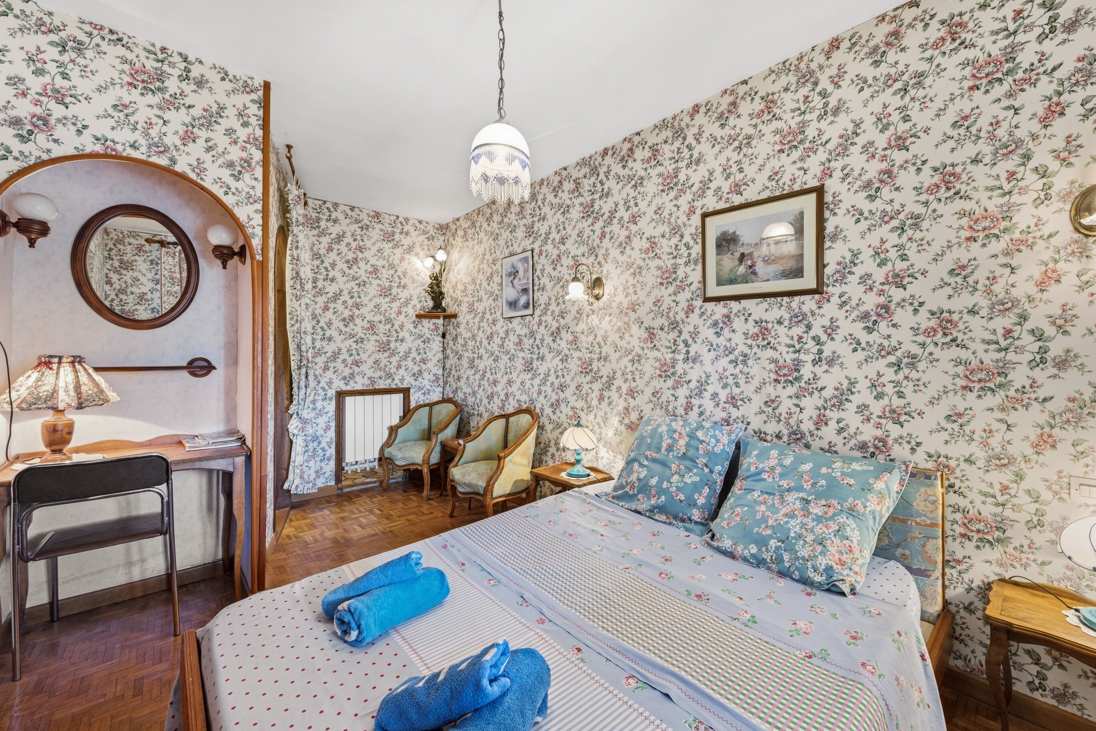 Une pièce avec un papier peint à motifs floraux, un lit avec une literie bleue à motifs floraux, un bureau avec une lampe et un lustre.