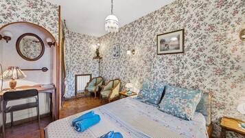 Een kamer met bloemenbehang, een bed met blauw bloemenbeddengoed, een bureau met een lamp en een kroonluchter.