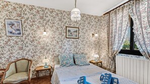 Une chambre avec un papier peint à motifs floraux, un lit avec une literie bleue et blanche, une chaise en bois, une table de chevet avec une lampe et une fenêtre avec des rideaux.