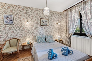 Un dormitorio con papel tapiz floral, una cama con ropa de cama azul y blanca, una silla de madera, una mesita de noche con lámpara y una ventana con cortinas.