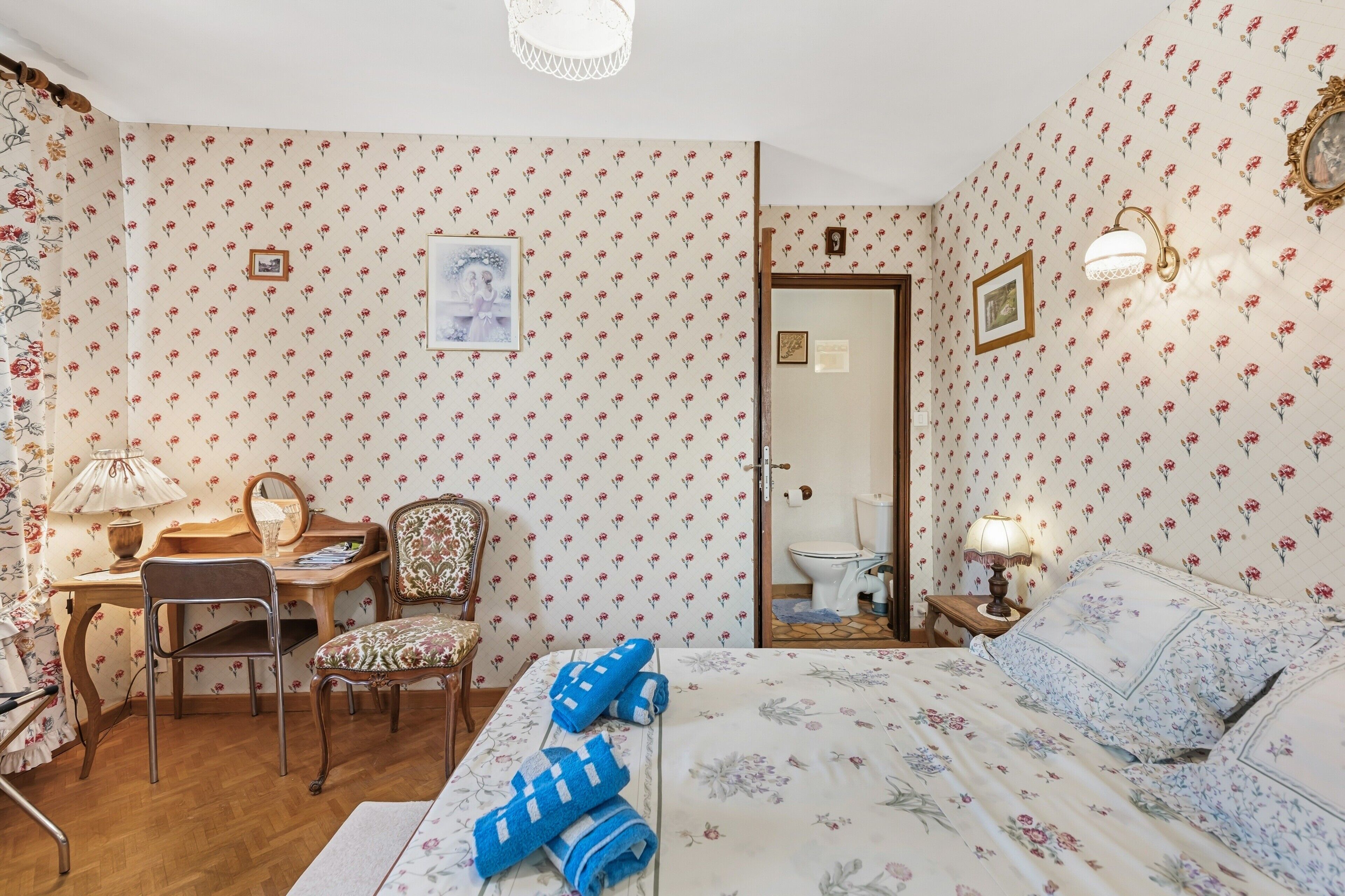 Ein Zimmer mit blumigem Wanddesign, einem Bett mit blauen Handtüchern, einem Holztisch mit Lampe, einem Stuhl und einem Spiegel.