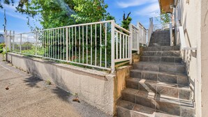 Un escalier en pierre mène à un bâtiment, lequel est entouré d’une clôture en métal blanc et est adossé à un décor de verdure.