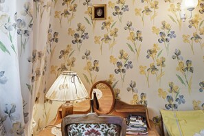 Une pièce dont les murs sont recouverts d’un papier peint à motifs floraux, avec une table d’appoint en bois sur laquelle se trouve une lampe, un tableau encadré et une chaise agrémentée d’un coussin à motifs.