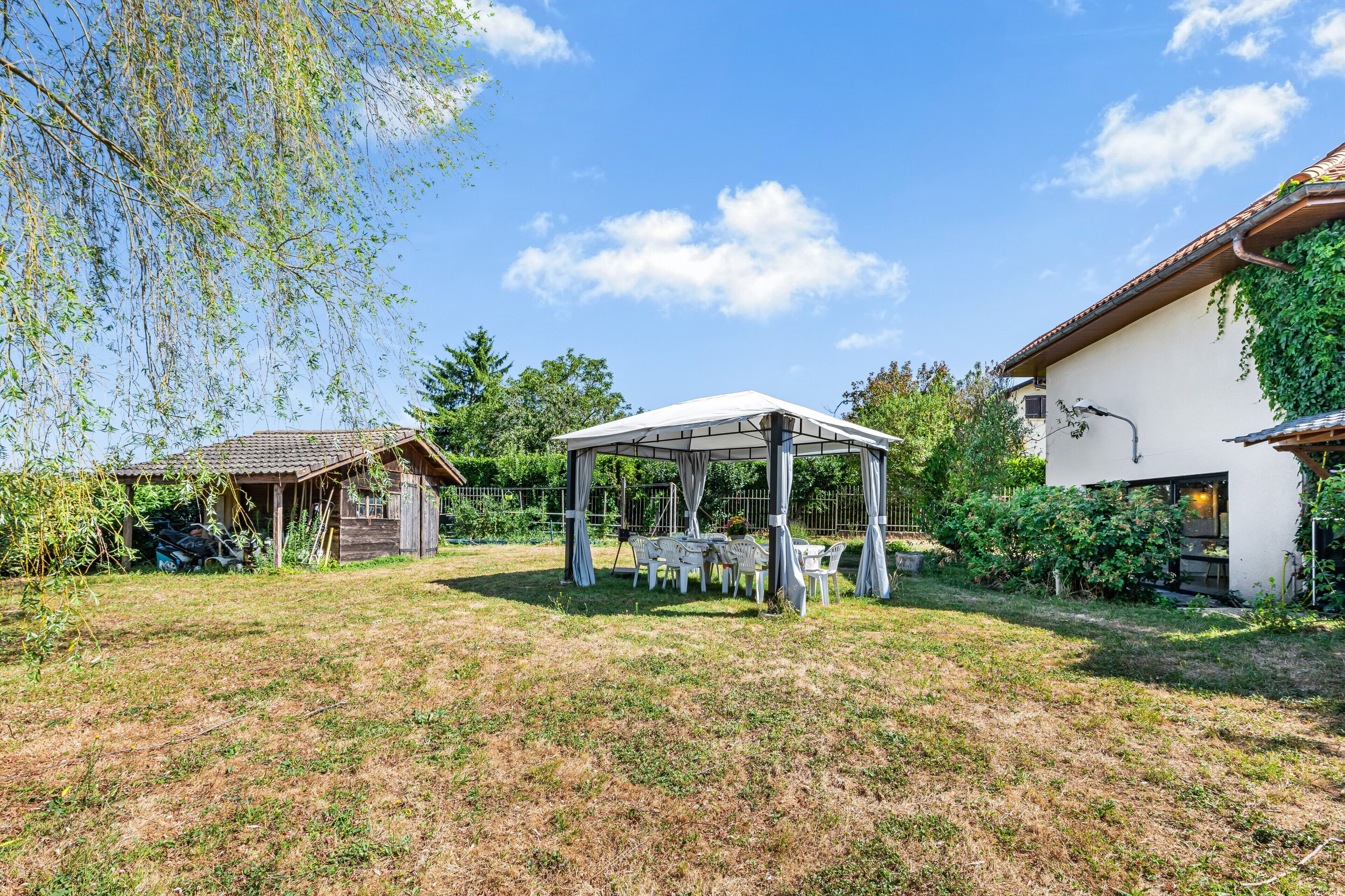 A covered outdoor seating area with tables and chairs, surrounded by a grassy lawn and a building with a tiled roof.