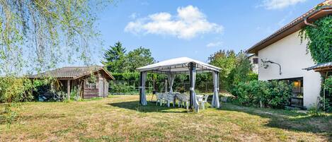 A covered outdoor seating area with tables and chairs, surrounded by a grassy lawn and a building with a tiled roof.