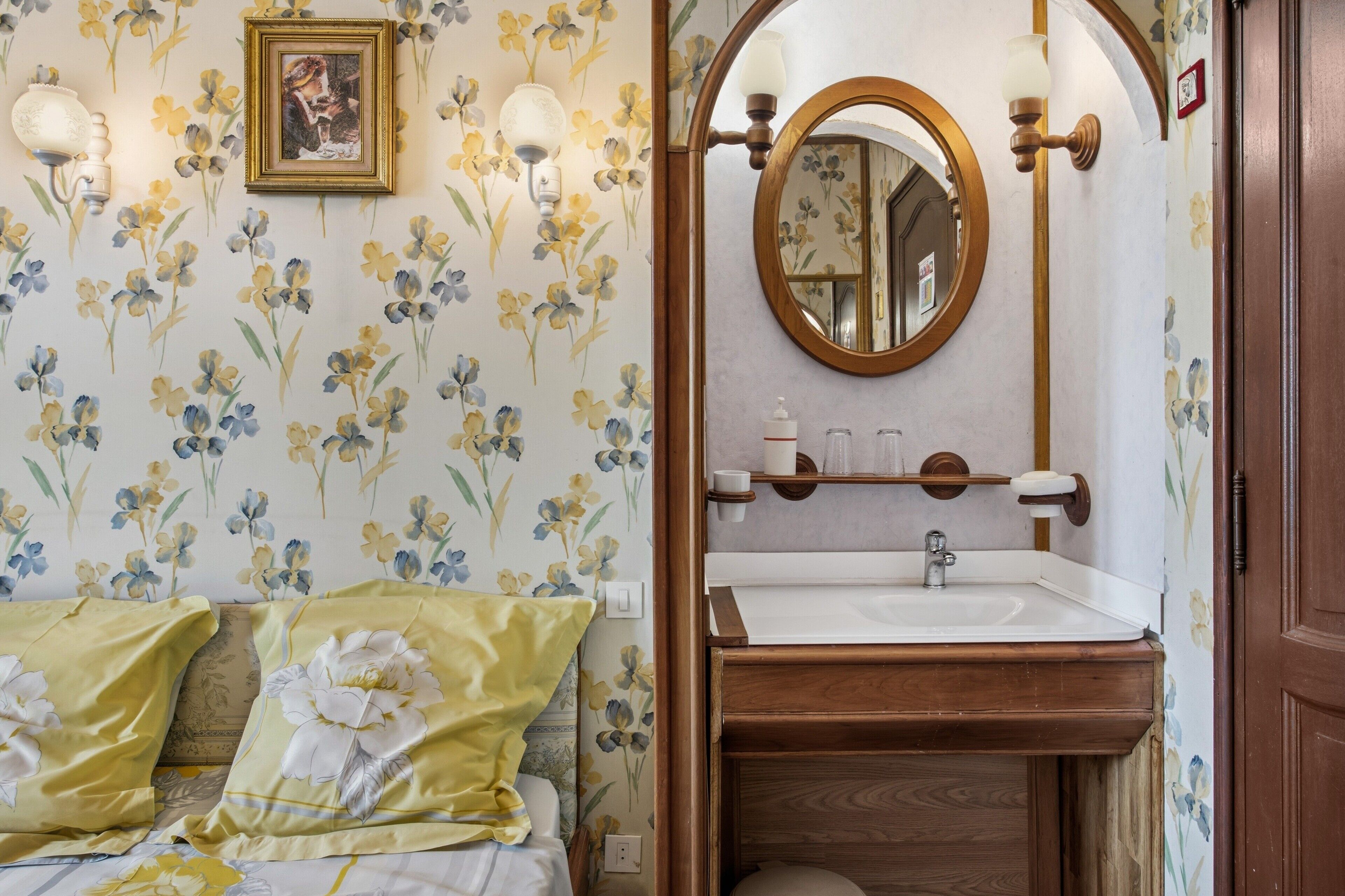 A bedroom with floral wallpaper, a bed with floral bedding, a bedside table, and a mirror above a wooden vanity with a sink.