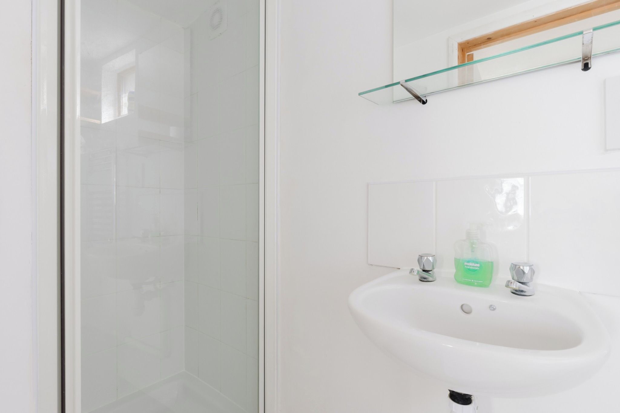 A bathroom with a glass shower enclosure, a white sink with two faucets, and a green bottle of hand soap.