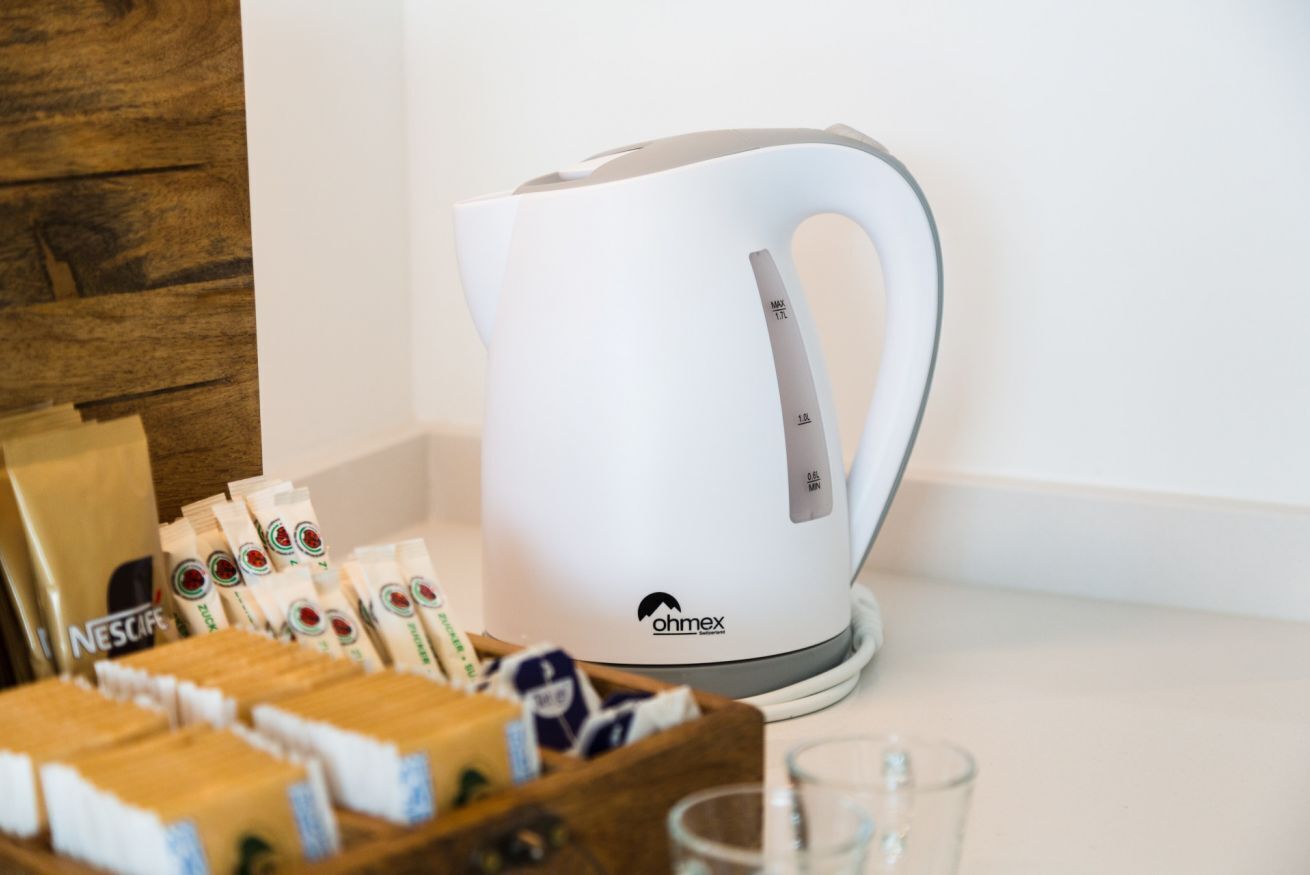 A white electric kettle with a handle, a wooden box with coffee pods, and a glass on a countertop.