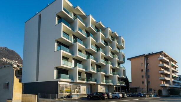 Un edifici de diverses plantes amb balcons, una zona d'aparcament i un cel serè.