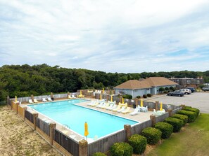 A rectangular swimming pool with a wooden fence, surrounded by lounge chairs and umbrellas, with a parking area and buildings in the background. - Kitty Hawk Family Oasis Pool & Tennis - 1 Mi to Beach (Kitty Hawk)
