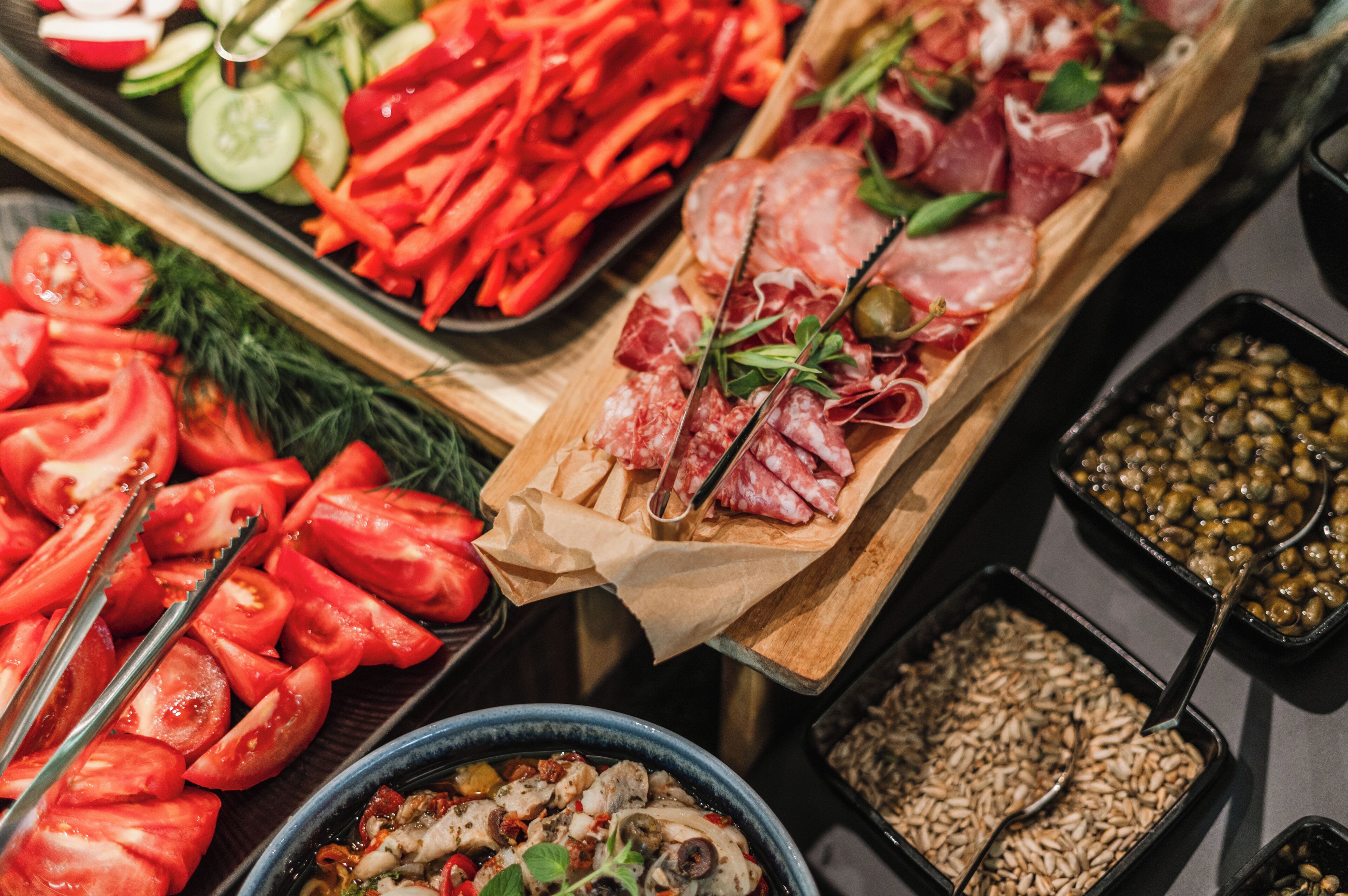 Diverse soorten eten, zoals plakjes tomaat, komkommer, rode paprika en verschillende vleessoorten, gepresenteerd op houten planken.