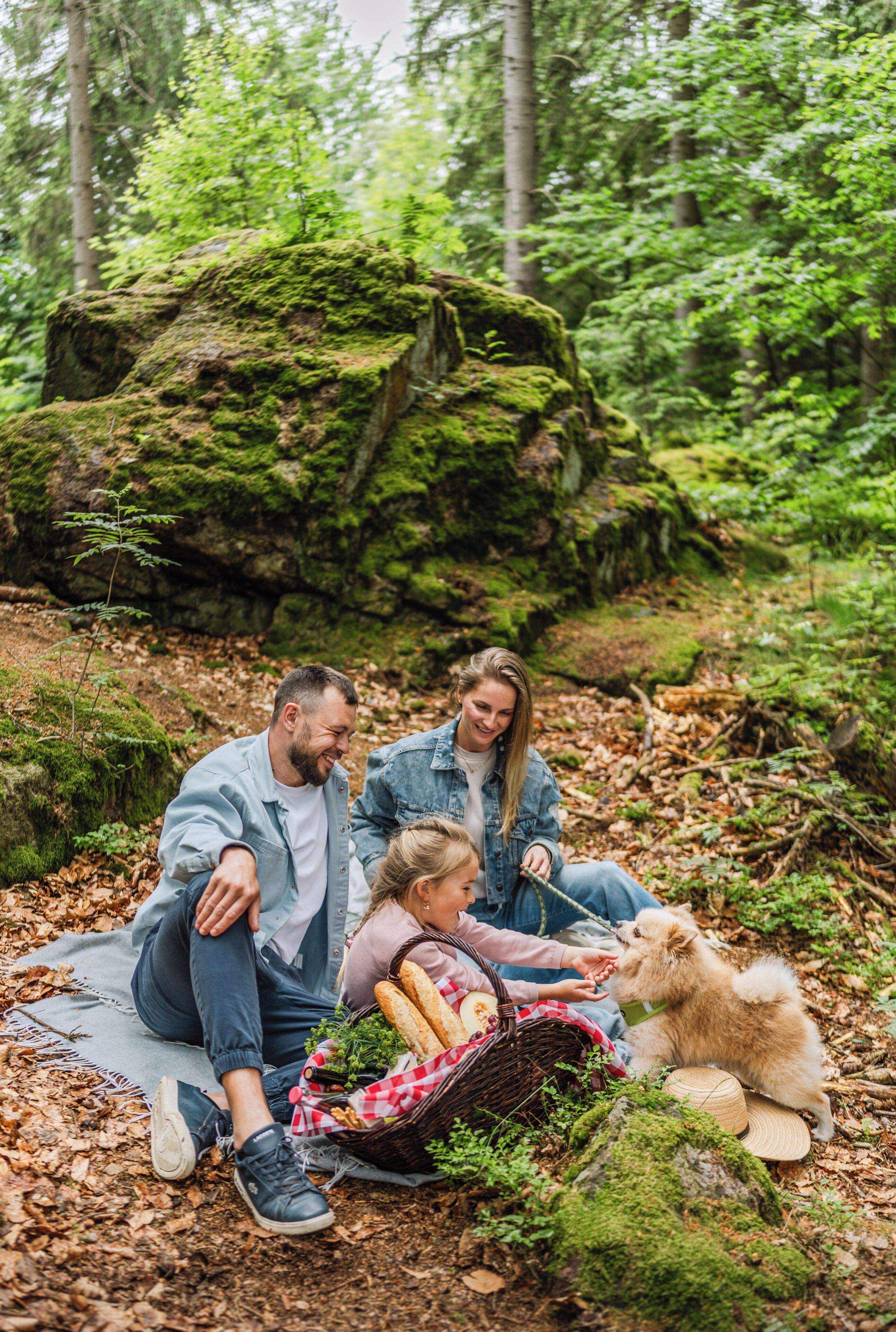 Una famiglia fa un picnic in una foresta, con una roccia ricoperta di muschio, una tovaglia da picnic e un cesto pieno di cibo.