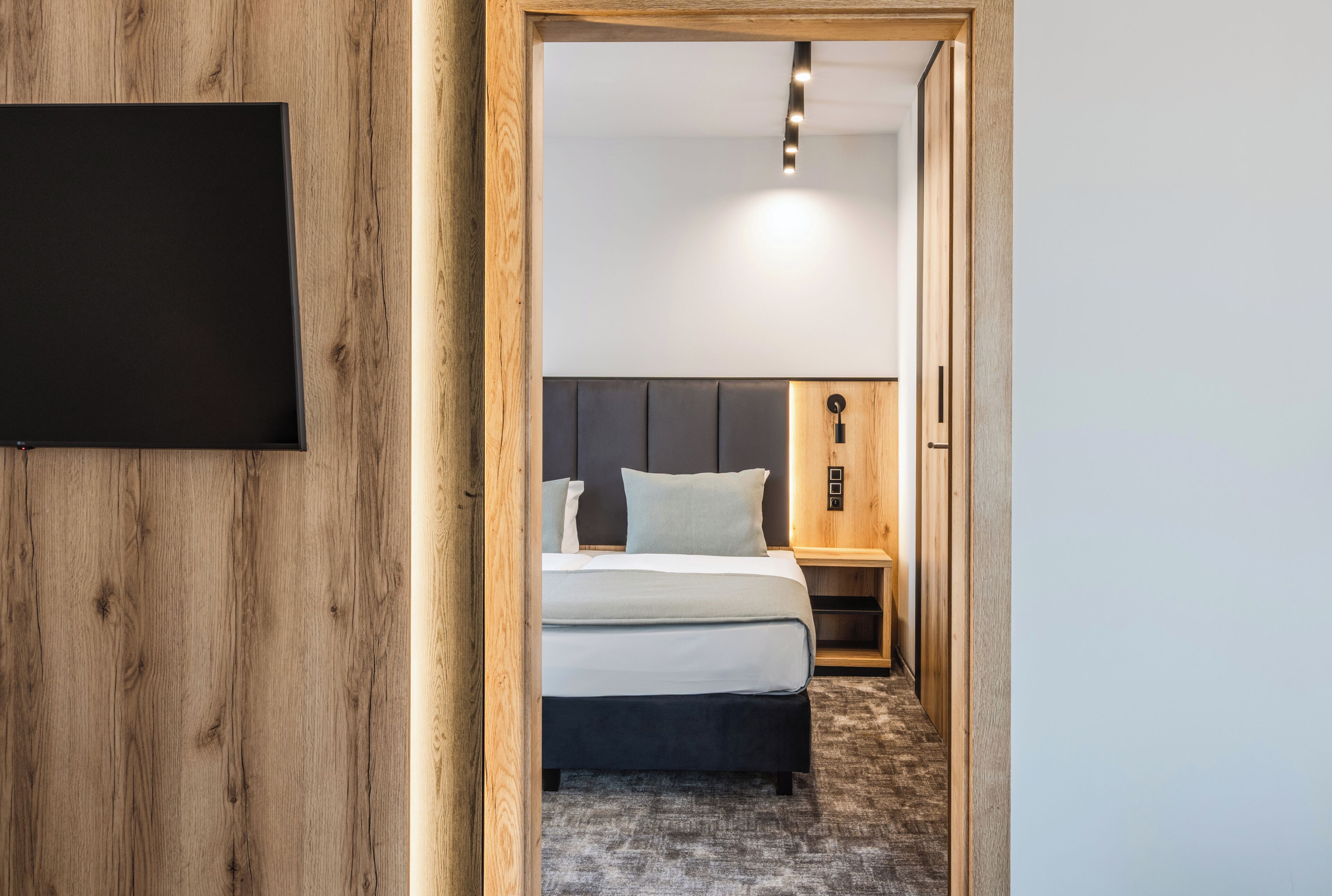 A hotel room with a wooden wall, a bed with a grey headboard, a bedside table, and a wall-mounted TV.