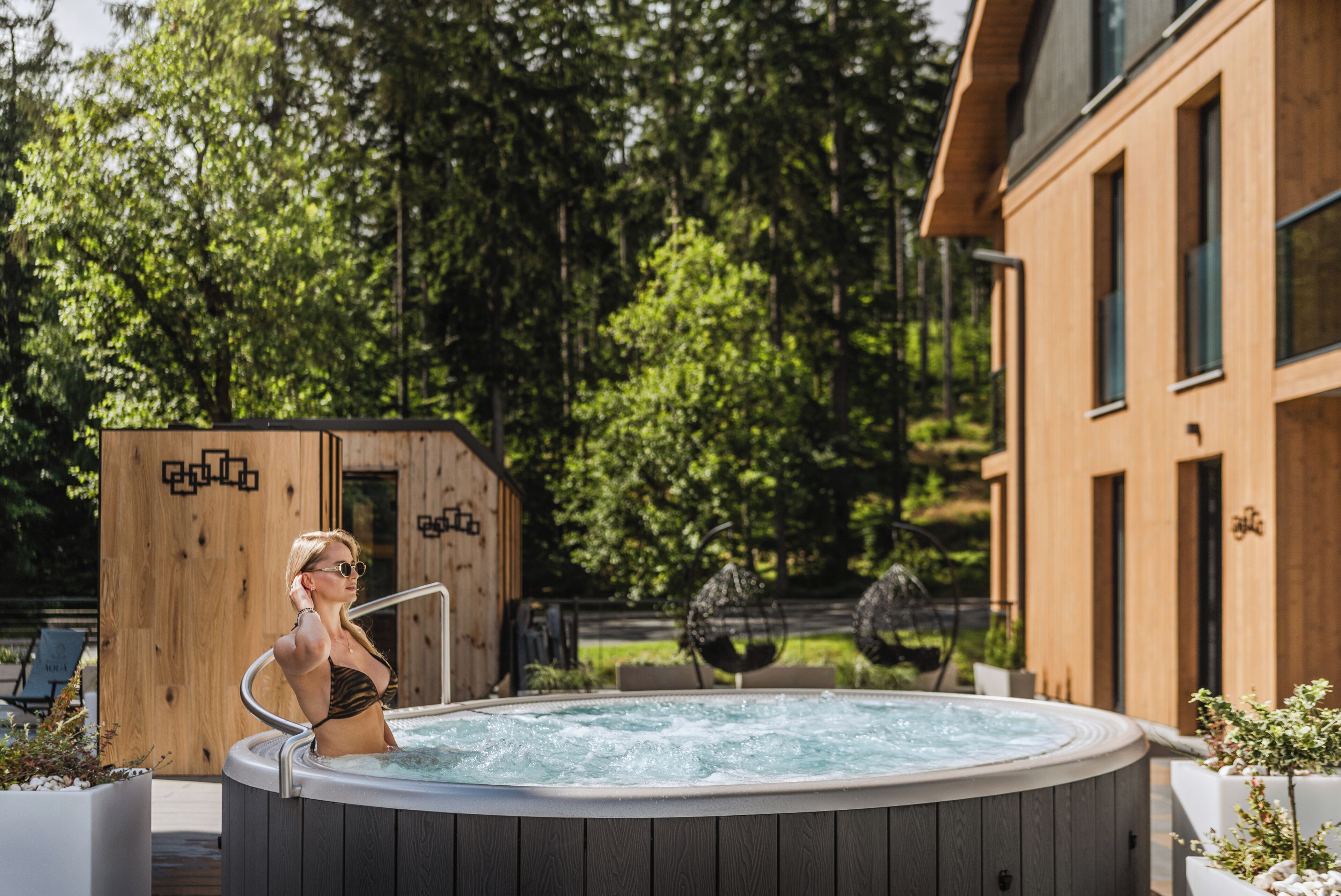 A person in a hot tub with a wooden deck and a building in the background.