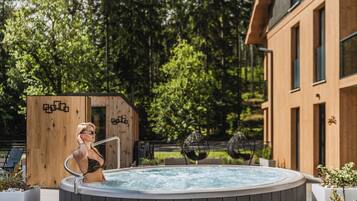 A person in a hot tub with a wooden deck and a building in the background.