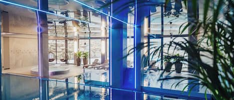 Una piscina interior con paredes de vidrio transparente, sillones de descanso y plantas en macetas.