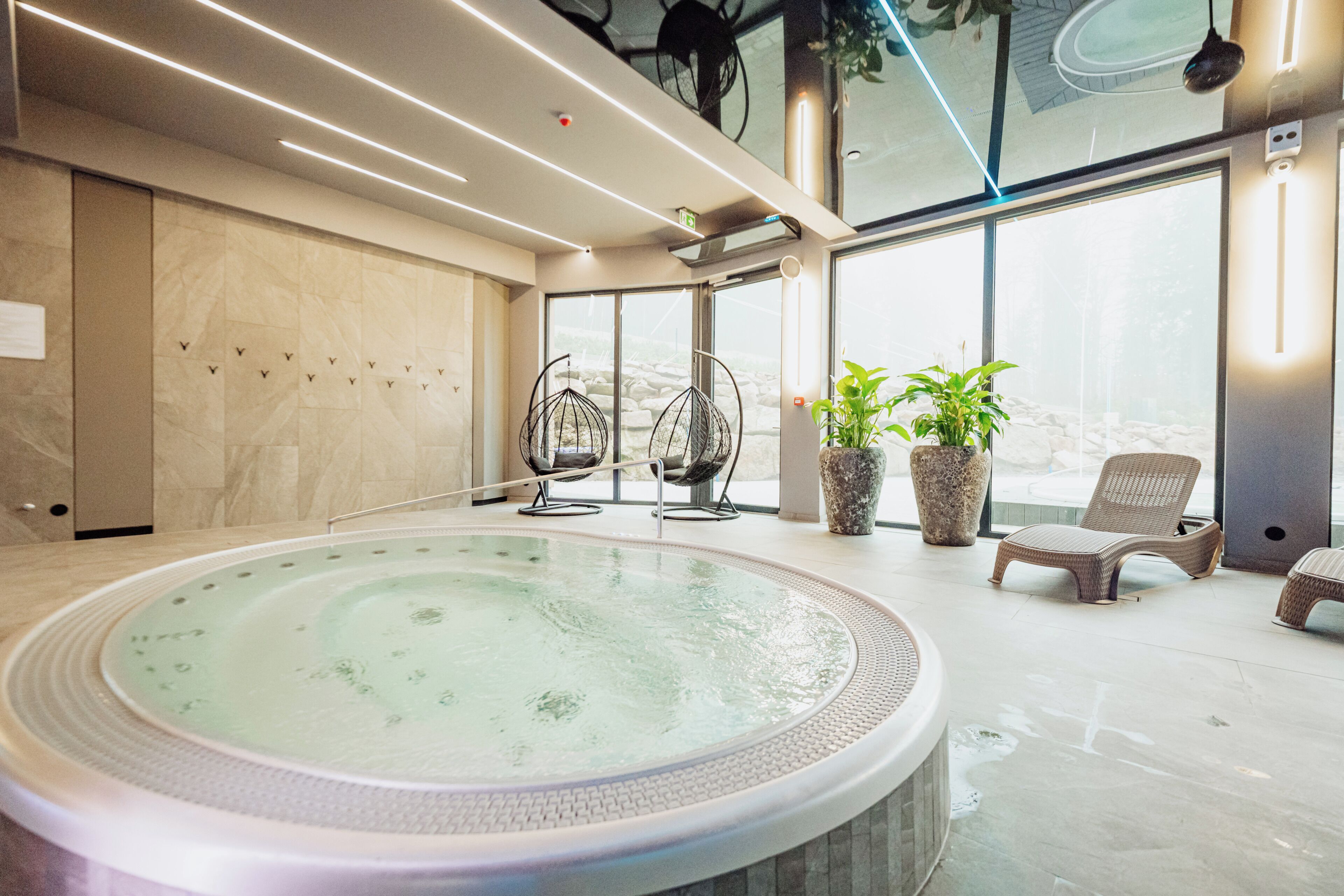Uma área moderna de piscina interna com hidromassagem, espreguiçadeiras e plantas em vasos.