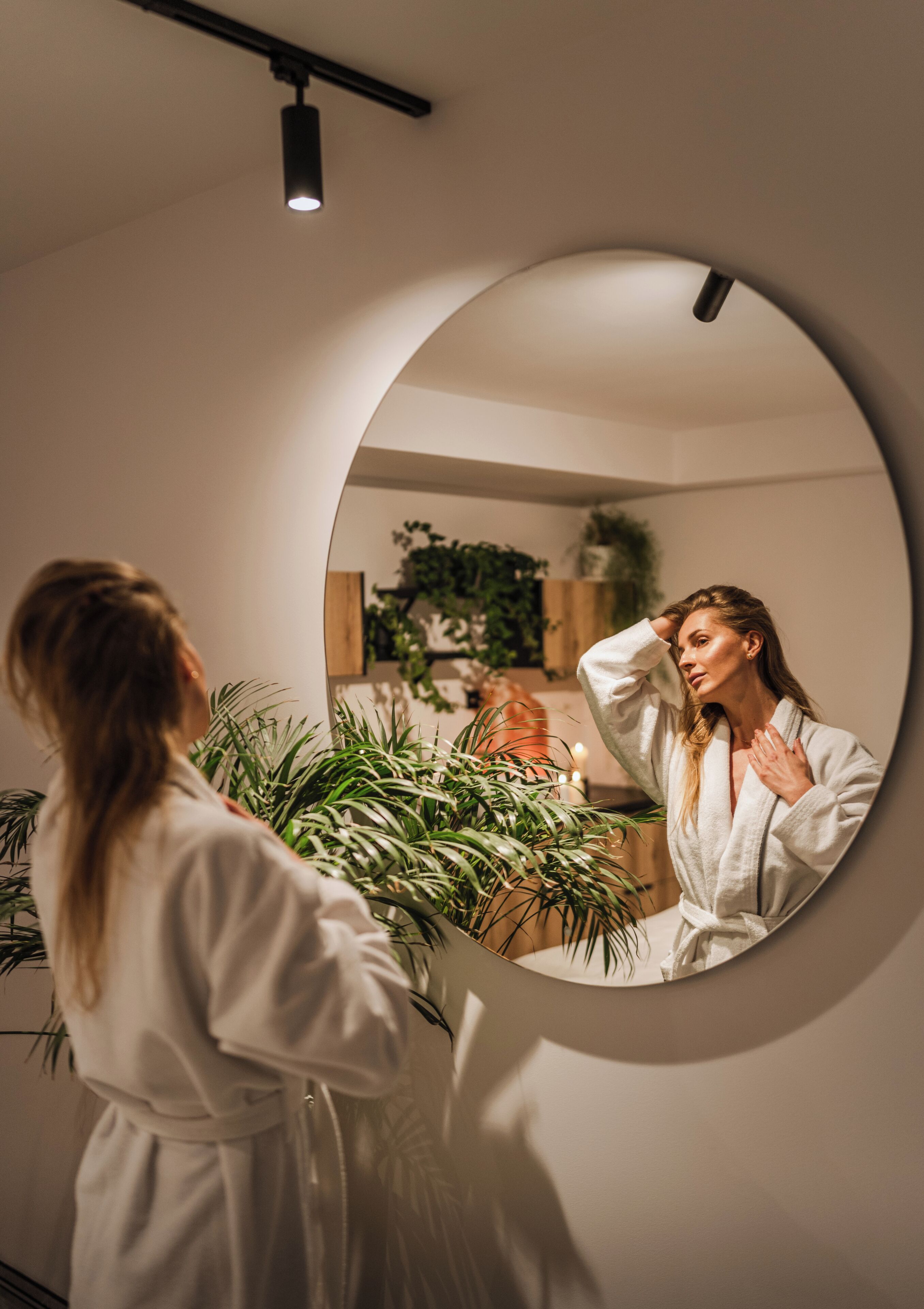 A person in a white robe standing in front of a mirror with a round frame, with a plant and a shelf in the background.
