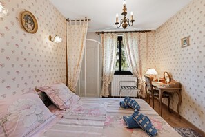 A bedroom with a bed, a desk, a chair, a window with curtains, a chandelier, and a radiator.