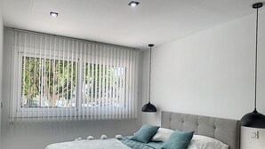 Um quarto com uma cama, mesa de cabeceira, janela com persianas e luminária pendente.