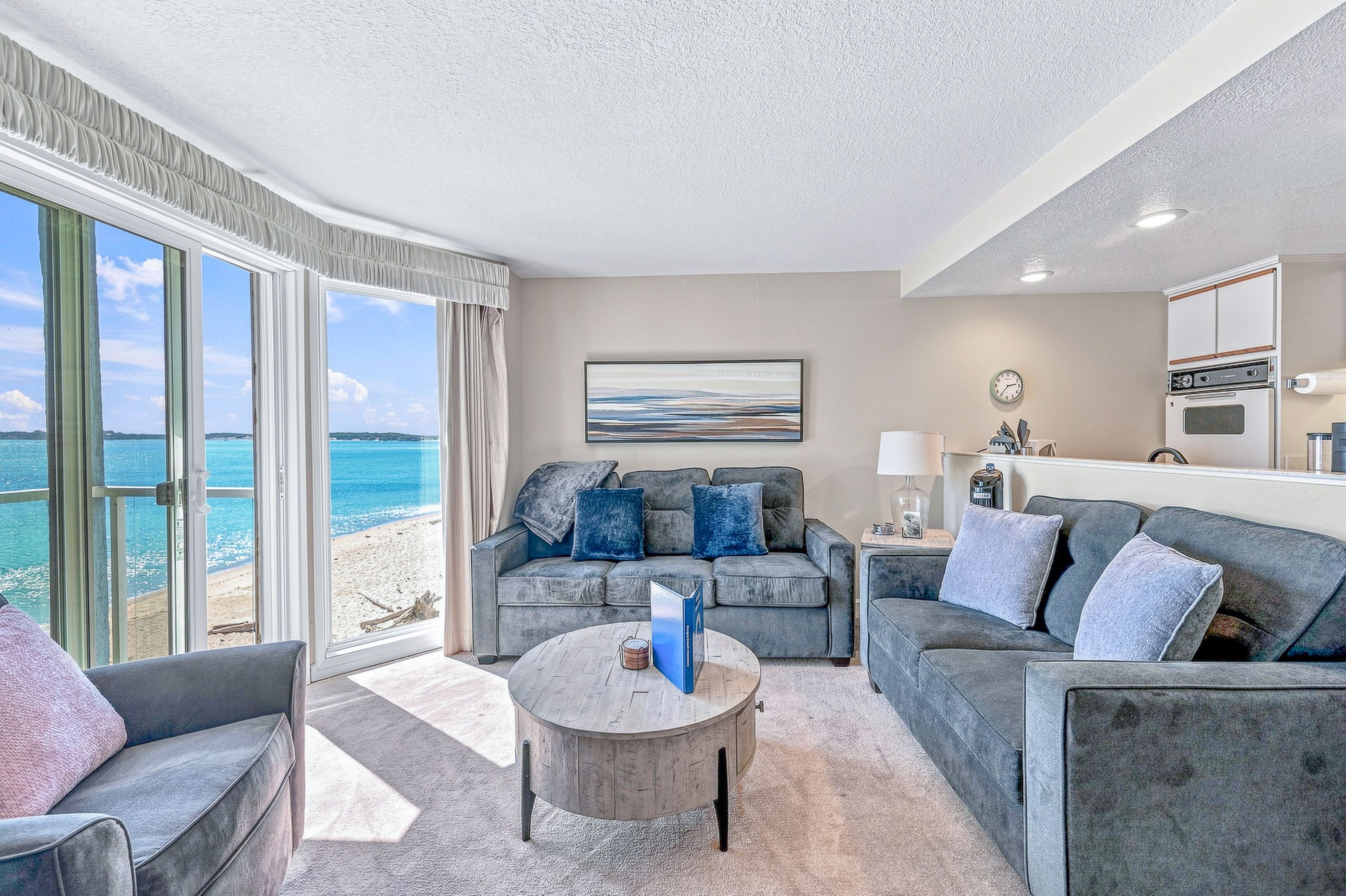 A modern living room with a view of the beach, a round coffee table, and a sofa with blue cushions.
