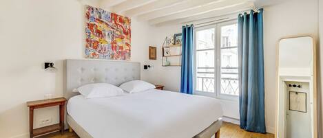Ein Schlafzimmer mit einem Bett, einem Nachttisch, einem Fenster mit Vorhängen, einem wandmontierten Spiegel und einem farbenfrohen abstrakten Gemälde.