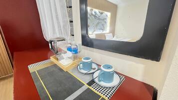 A hotel room with a bed, a bedside table with a kettle, a bottle of water, and a blue mug.