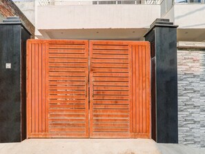 Ein geschlossenes Holztor mit vertikalen Lamellen, flankiert von schwarzen Säulen, führt zu einem Gebäude mit weißer Fassade und einer Steinsäule.
