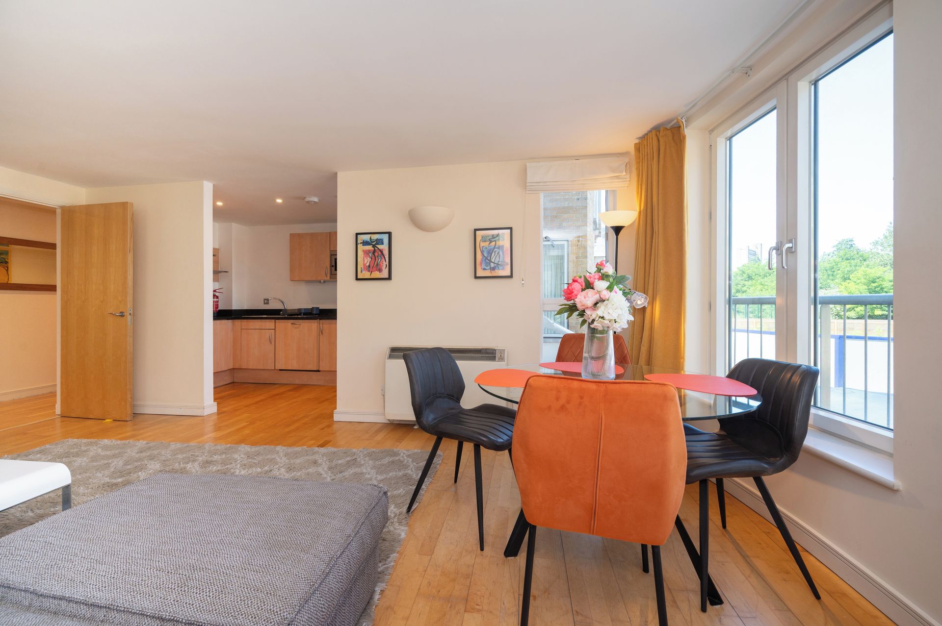 A modern living room with a dining area, a kitchen in the background, and large windows with curtains.