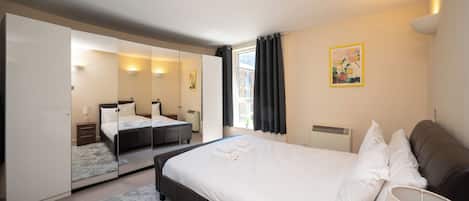 Une chambre d’hôtel avec un grand lit, un miroir, une table de chevet, une lampe, une fenêtre avec des rideaux et un tableau au mur.