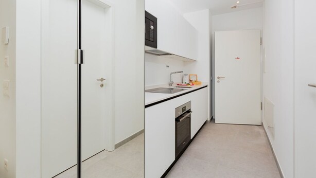 A narrow hallway with white walls, a kitchen area with a sink, stove, and oven, and a door with a fire alarm. - {Suite 501} Gym, Business Room, Modern, Relax (Lugano)