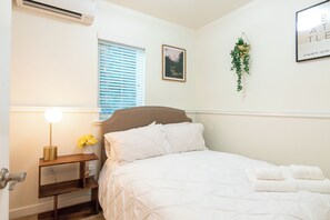 A bedroom with a bed, bedside lamp, air conditioning unit, window with blinds, framed picture, and a wall-mounted plant. - Seattle 2 Bedroom 1 Bath Suite (Seattle)