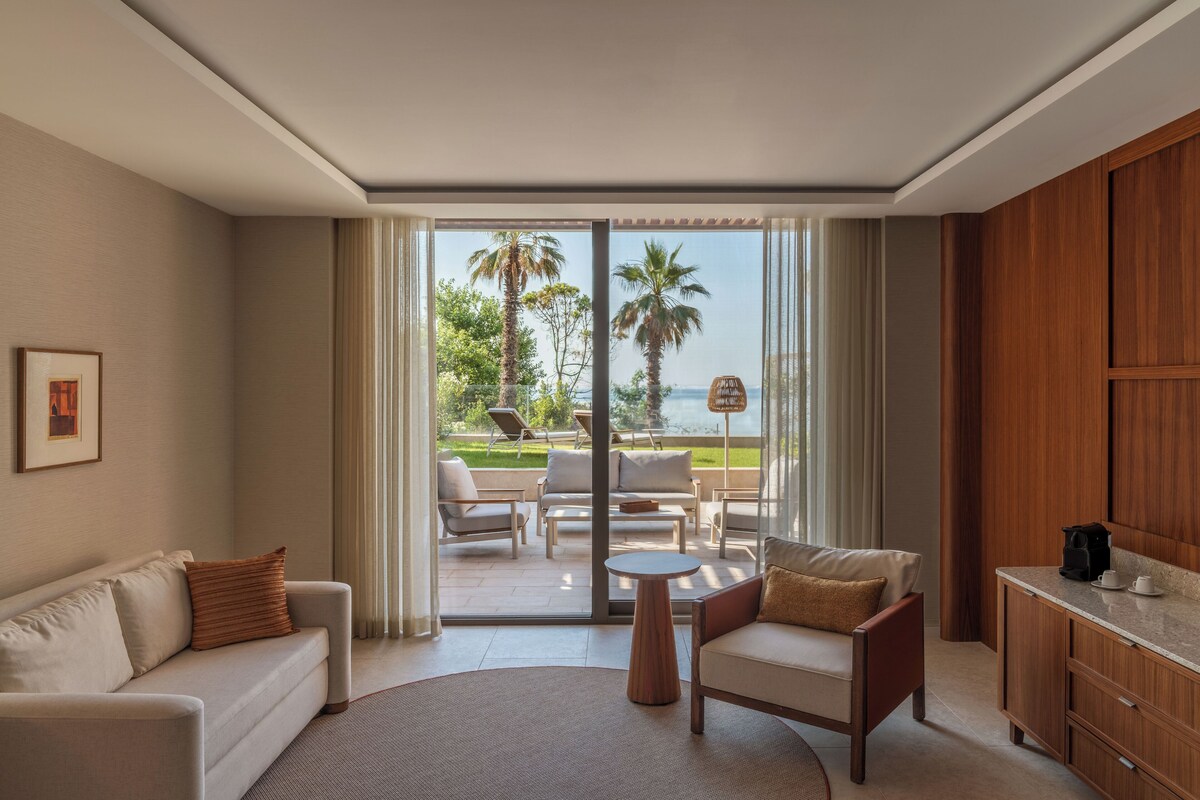 Habitación de hotel con sofá, sillón, una mesita redonda y vistas al exterior a través de amplias puertas correderas.