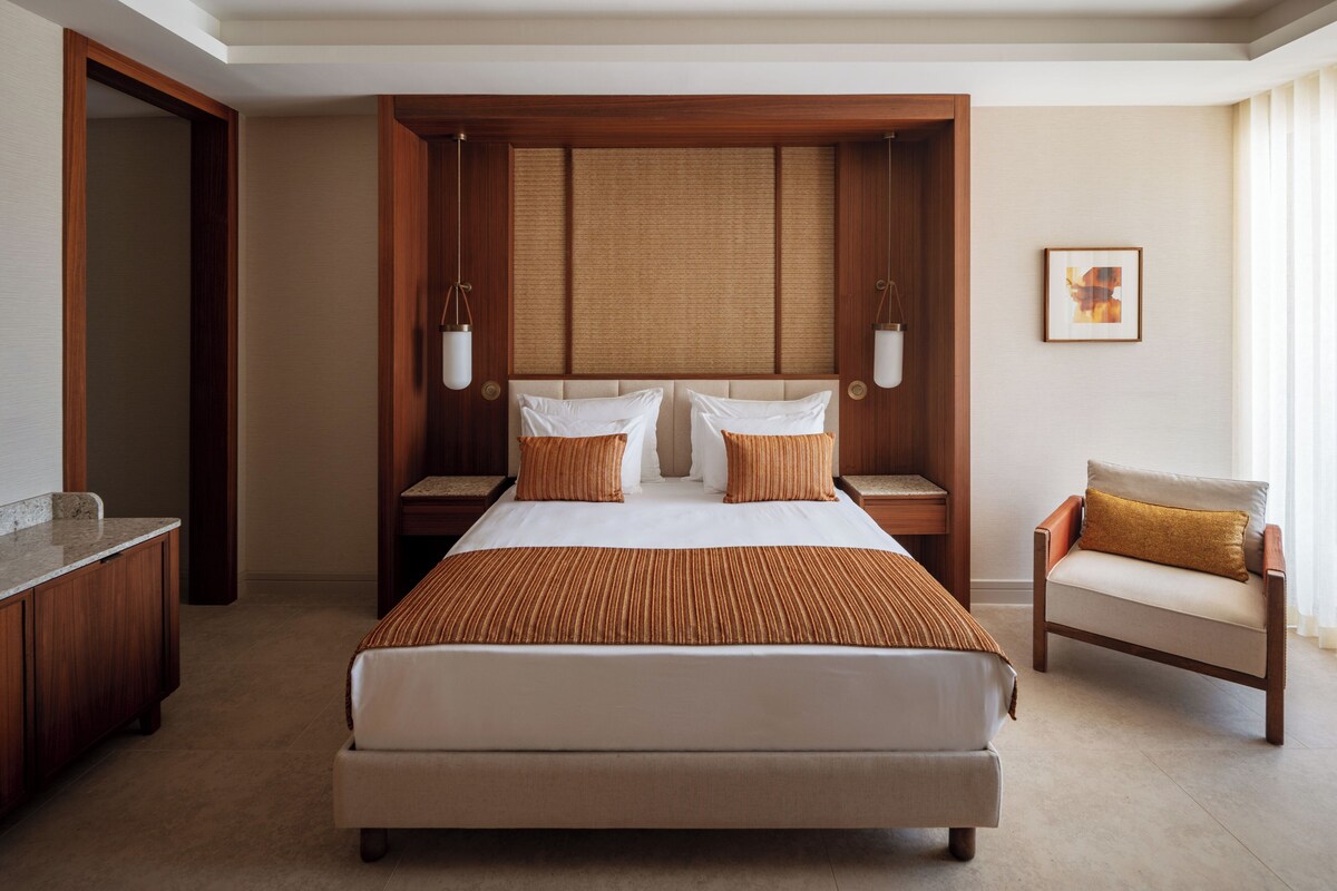 Habitación de hotel con una cama grande, cabecera de madera, dos mesitas de noche, una silla y un cuadro en la pared.