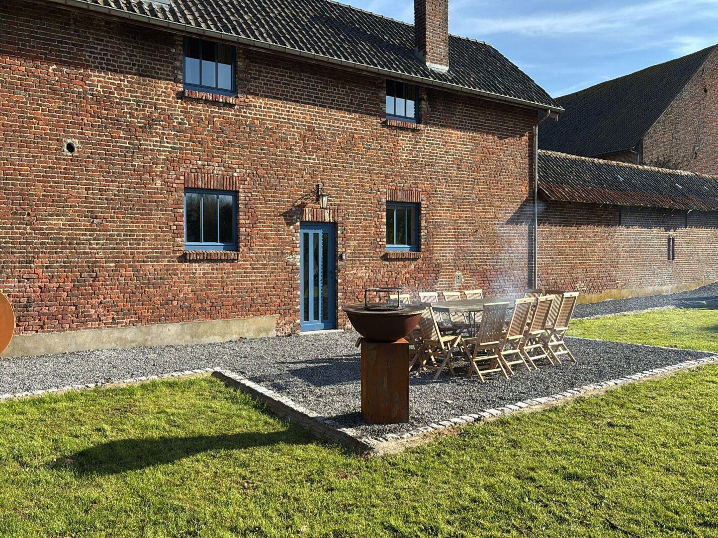 A brick building with a blue door and windows, a patio with a table and chairs, and a fire pit.