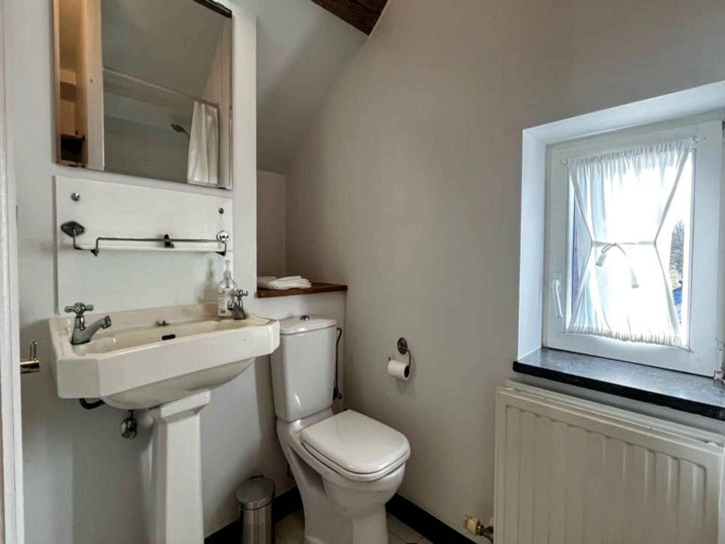 Une salle de bain avec un lavabo blanc, un miroir et des toilettes. On y trouve une fenêtre avec des rideaux, un radiateur et un porte-serviettes.