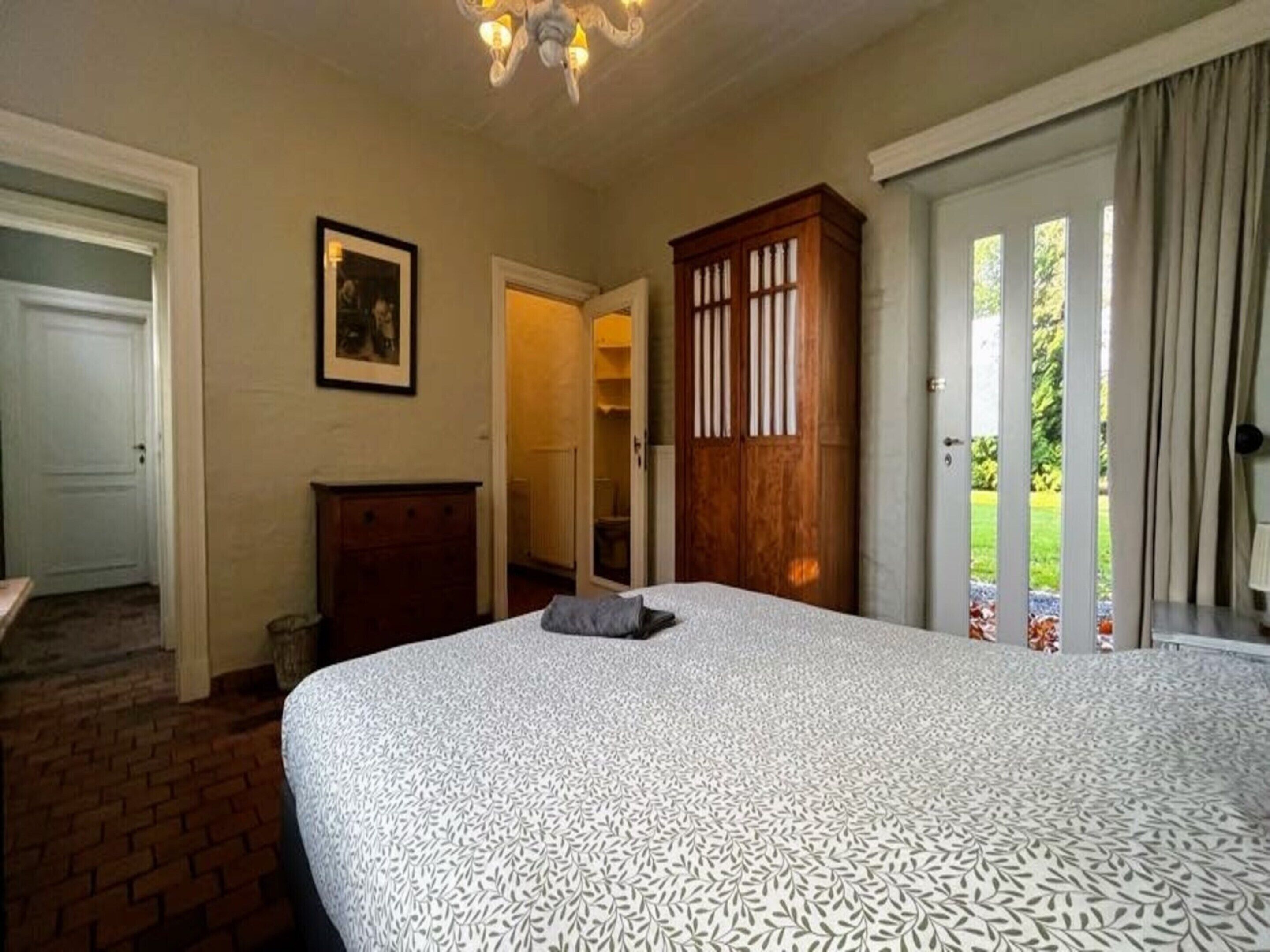 A bedroom with a large bed, a wooden wardrobe, a door leading to a balcony, and a framed picture on the wall.