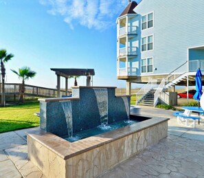 A water feature with a tiered design, a covered seating area, and a building with multiple balconies in the background. - Serene 2-bedroom condo with AC in charming Galveston (Galveston)