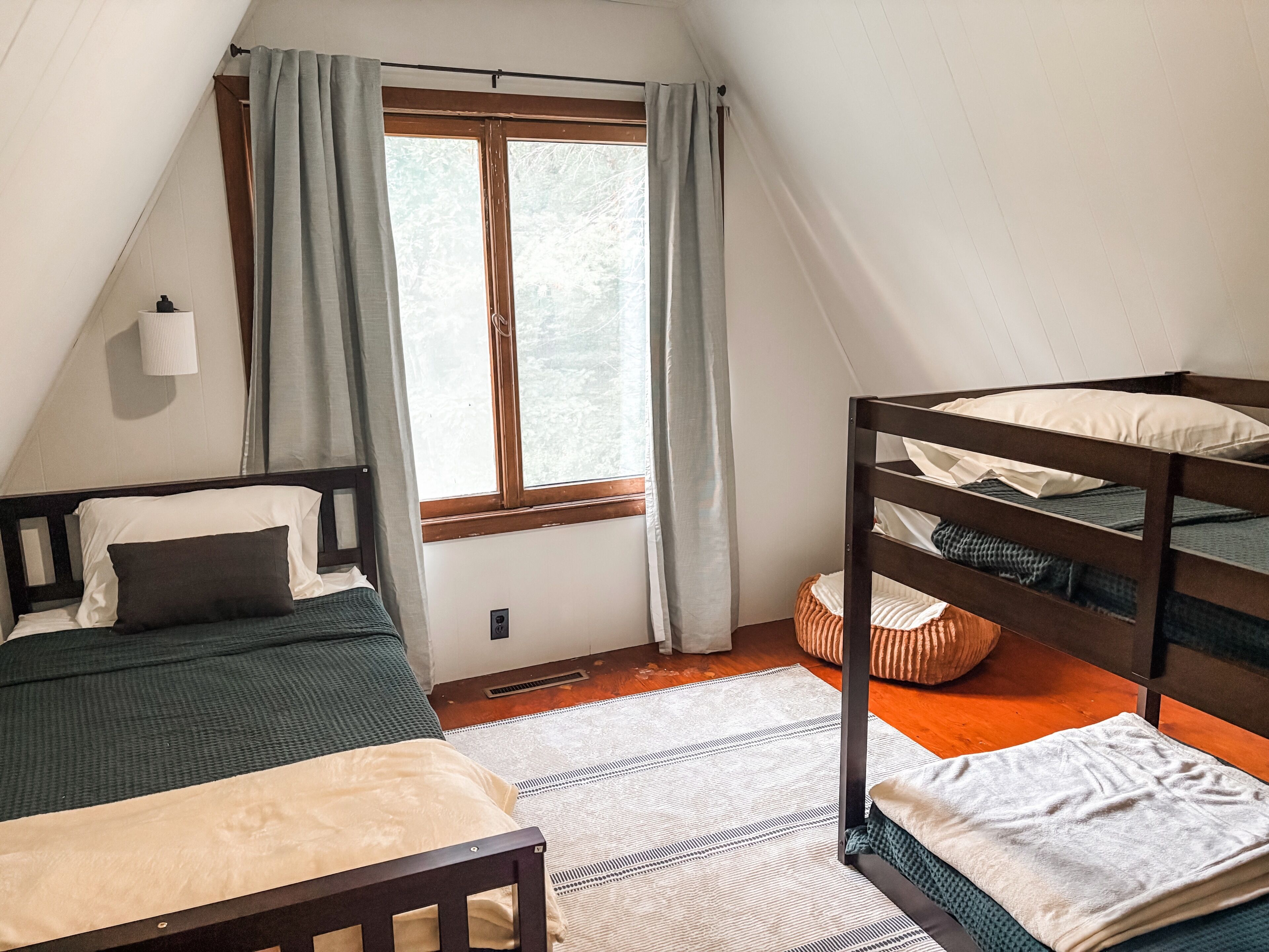 A room with a bunk bed, a single bed, a window with curtains, a lamp, and a rug.